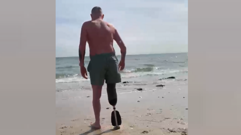 Alex Young walks away from the camera on a sandy beach towards the sea. He wears swimming shorts and a prosthetic right limb.