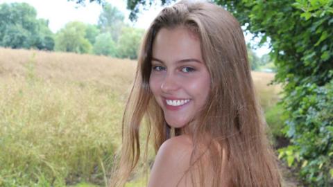 A woman has blonde/mousey hair. She is turning to face the camera. In front of her is a field to the left and some trees to the right