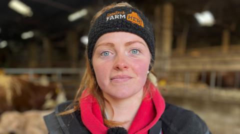 Young farmer Nicola Wordie stands in a cattle shed wearing a black headband, red hoodie and black jacket.