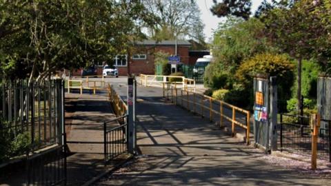 A path with railings on either side leading to a single-storey brick school building. There are trees and shrubs lining the front gates of the premises