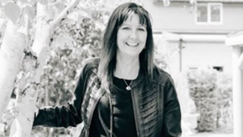 A black and white image of a smiling woman with shoulder-length hair wearing a leather jacket and necklace
