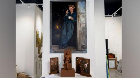 A large painting of a female steelworker leaning against a door frame is attached to a wall inside Sunderland's 24NE Gallery. The painting is in dark colours and shows the woman holding a cup in one hand a cigarette in another. The statue maquette is on a table beneath the painting. It is bronze-coloured and shows a female steelworker sitting with her legs crossed.