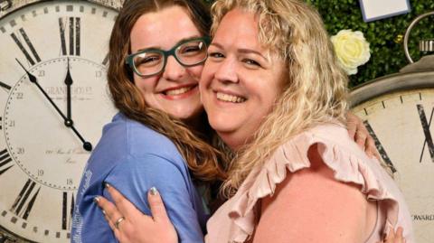 Two women hugging and smiling. The woman on the left has long dark hair, glasses and is wearing a blue T-shirt. The woman on the right has long blonde hair and is wearing a light pink top. They are stood in front of two large old-fashioned clock faces.