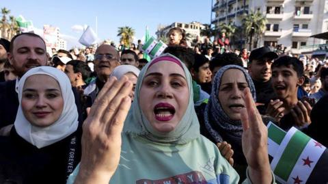 A woman gestures at the camera, with a crowd of other people celebrating behind her