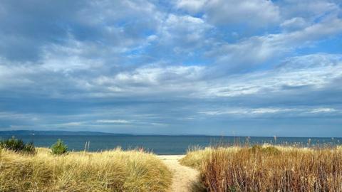 Cloudy blue sky above darker blue sea with sand and sand dunes in the foreground