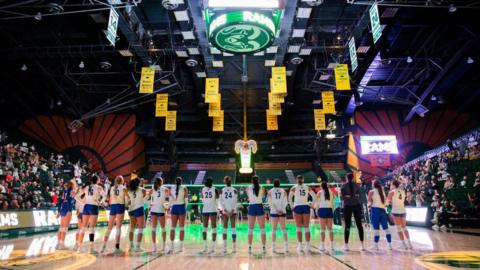 San Jose State University Spartans players line up to sing the national anthem at the NCAA Mountain West women's volleyball game against the Colorado State University Rams in Fort Collins, Colorado on 3 October. They wear white vests and blue shorts. Spectators are sitting on each side of the court.