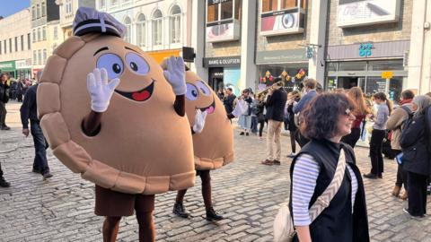 Two people dressed in giant past costumes waving their arms in the air as they walk through a street in Truro