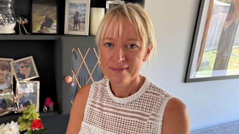 Tracy Venison has blonde hair and is smiling. She is wearing a white top and is sat in front of photos on a cabinet