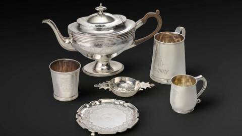 Silver items including a plate, jug, and three cups, against a dark background.