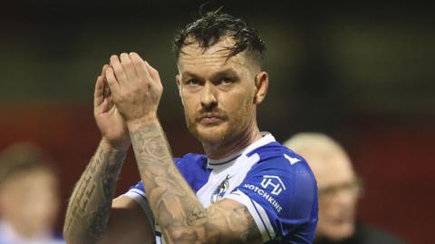 Josh McEachran stands on the pitch and holds his hands up and claps. He is wearing Bristol Rovers kit