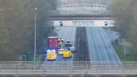 Police cars and ambulances block the road. There are several cars and a red lorry on the road. There is a banner on a bridge above the road reading Port of Felixstowe.