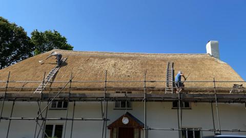 Two men work on an almost finished thatched roof against a bright blue sky. 