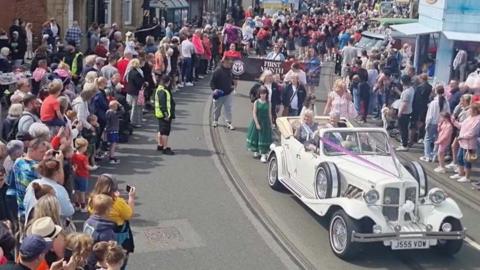 A vintage white car driving through crowds in Fleetwood