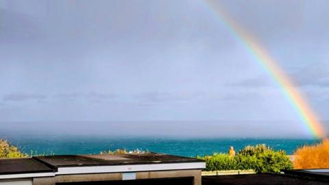 A rainbow arches into the grey sky on right side with green/blue sea below