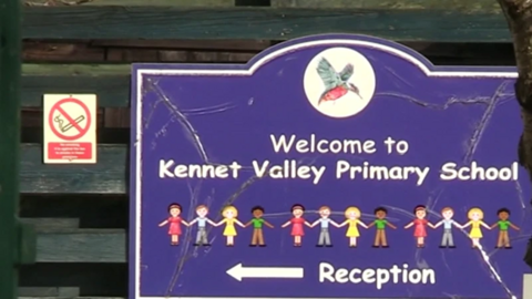 A purple sign for a school. It has a picture of a bird at the top, the words Welcome to Kennet Valley Primary School and cartoon drawings of children below. At the very bottom of the sign, there is an arrow pointing towards the reception. There is a no smoking sign in the top left of the image.