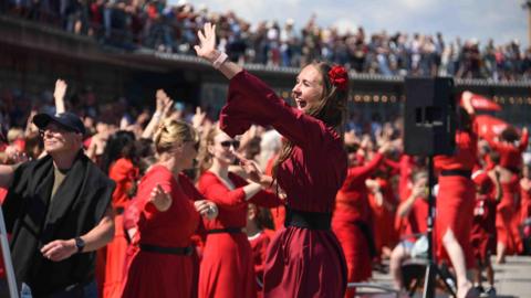 A woman, wearing a red dress and headband, leads a group of dancers, also dressed in red, to a choreographed routine to Kate Bush's Wuthering Heights.