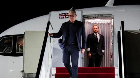Sir Keir Starmer walks down a staircase from an aircraft at night. He is wearing a dark blue suit and a black shirt. He has short grey hair and wears black spectacles. He has a serious expression. A cabin member is visible inside the plane through the open door, watching on with one hand in his pocket.