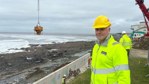 Rob Braddick is wearing a yellow hard hat and a yellow hi-vis jacket. A yellow container holding concrete is on the swing arm of the crane over the beach behind him.