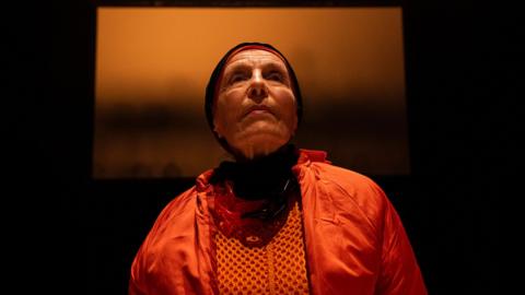 A woman wearing orange clothing and black hood looking out in the distance. The background is dark with a large orange picture.