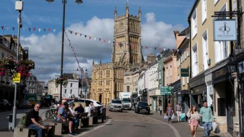 A road in central Cirencester with shoppers and bunting, leading up to the Parish Church of St John Baptist at Market Place
