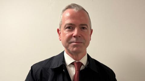 Pól Deeds has short grey hair swept back from his forehead and is wearing a dark coat, with a white shirt and red tie