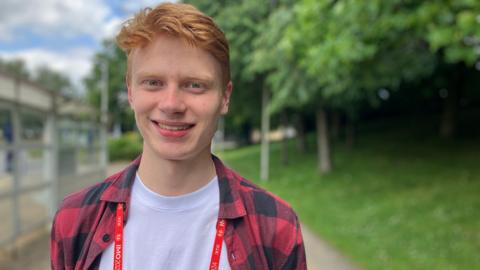 Albert Ben Carlson. He has red hair and blue eyes. He is wearing a white t-shirt with a red and black check shirt on the top. He is looking directly at the camera and smiling.