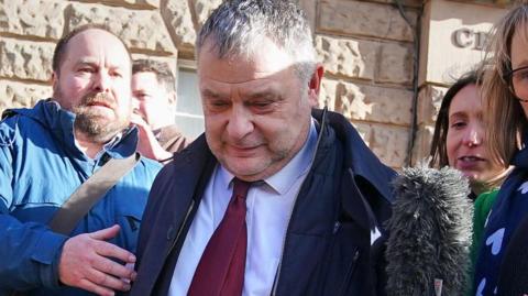 Mike Amesbury, a middle-aged white man with short, grey hair, is wearing a black jacket with a white shirt and dark red tie. On top of his suit he wears an unzipped dark coat. He is surrounded by journalists, and there is a large fluffy microphone being pointed towards him.