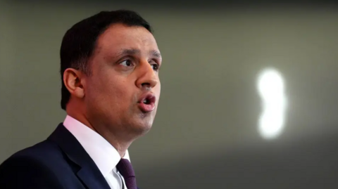 Anas Sarwar in a dark suit with a white shirt and purple tie