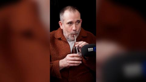 Xand van Tulleken blowing into a glass of water with a straw. He's wearing an orange jacket.