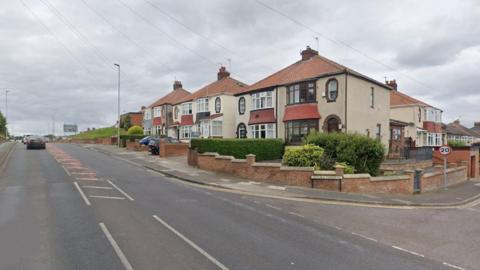 The junction of Ryehill Gardens and Hart Lane. Cars are driving up the road away from the camera on Ryehill Gardens. The road is lined with semi-detached houses.