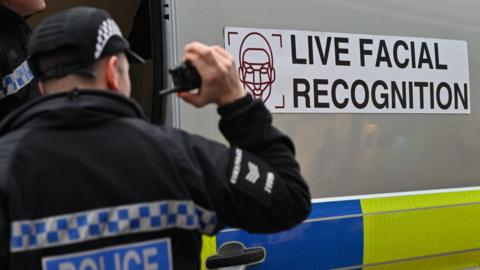 Police officer holding walkie-talkie stands by a police fan marked "Live Facial Recognition"