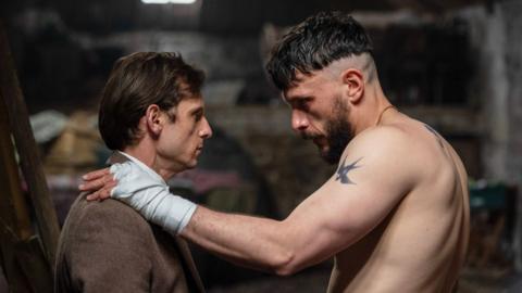 Richard Gadd faces Jamie Bell and puts a hand on his left shoulder. Richard Gadd isn't wearing a top whilst Jamie Bell is in a full suit.