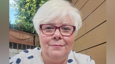 Jane Bailey, wearing glasses and a white spotted blouse, in a garden 