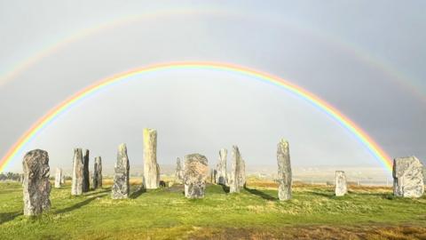 A view of the Calanais Standing Stones on the Isle of Lewis. The stones are mainly grey with light patches on a green grass field. The background is grey. A double rainbow is overhead. The lower rainbow is bright. The upper rainbow is fainter.
