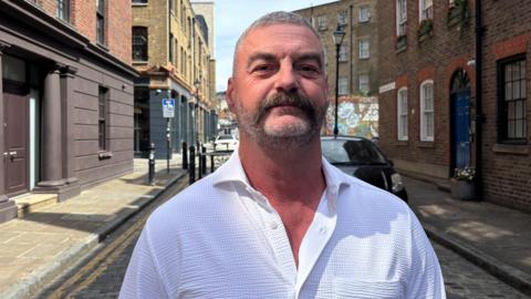 Chris Lloyd in a white shirt. He has short white and grey hair and a short beard. Elder Street is in the background, with brick buildings and a parked car.