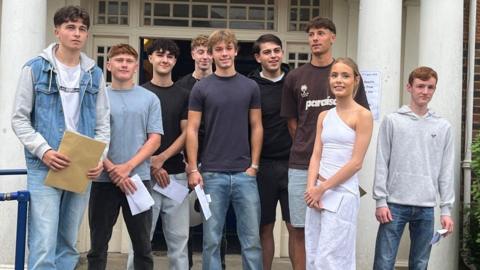 Eight young men and one young woman stand smiling on the front steps outside Ashcombe School library where they collected their A-Level results 