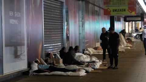 Several people are sleeping rough on the pavement under a building's awning. An advert saying "UNLEASH" hangs above them and the shop's shutters are down. 