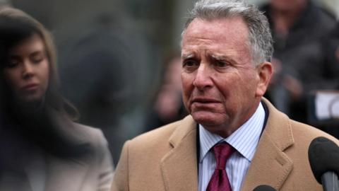 Witkoff in tan coat, red tie in front of woman in gray coat and next to black microphone