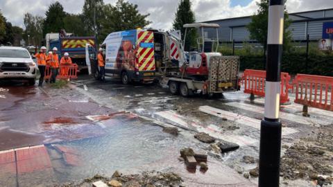 The image shows a burst water main on a zebra crossing on Lyde Road in Yeovil. There is water all over the road. Five men in orange high vis coats and white hard hats are surveying the damage.