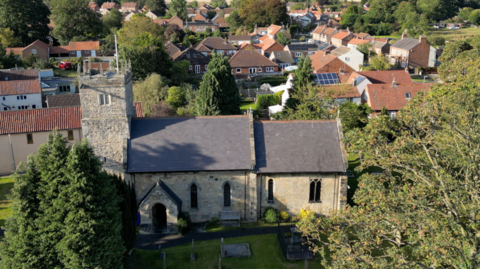 A birds eye view of Shiptonthorpe, focusing on the church with several houses huddled amongst it. The trees nearby cast a shadow on the stone walls of the church.