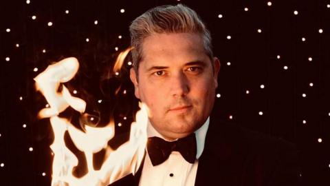 A man looking towards the camera wearing a bowtie and a black suit. There is fire on the left, and he is standing in front of a black background.