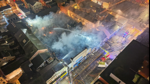 An aerial view of he building on fire with a fire engine spraying water into the area amid smoke and orange fire in one part of the building