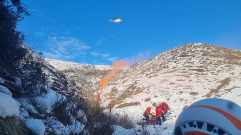 A helicopter approaching the scene of a rescue in a snowy mountain valley, with a smoke flare guiding the way in