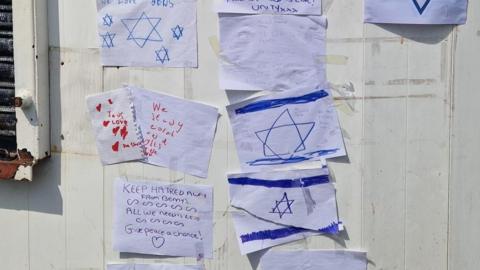 A white wall with a number of pieces of paper with handwritten messages and stars of David flags on them. 