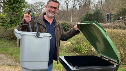 Jon Neal is standing on a gravel drive with trees and bushes in the background. He is holding a small grey caddy in one hand while standing next to a black and green wheelie bin, which he is holding open. He is wearing blue jeans, a blue jumper and a brown jacket. He is wearing glasses.