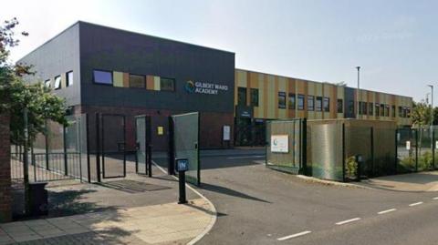 Gilbert Ward Academy, a modern school building with brown and yellow cladding. There is a tarmac entrance and a metal fence.