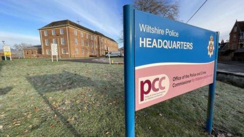 The Wiltshire Police Headquarters on a frosty morning. There is a large rectangular brick building in the background and a blue sign in the foreground. The sky is a pale blue with wispy clouds, and the trees are bare of leaves.