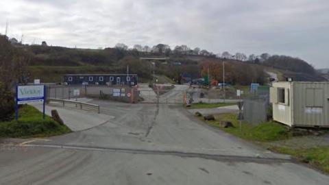 The entrance of a quarry landfill site. A portacabin is on the right hand side and a sign on the left that says Viridor. A gate is closed across a road in the centre. In the background there in a hill with bushes and trees.  