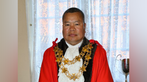 A nepalese man in a red mayor's cloak and white crevette, as well as gold mayoral chains. He has a shaved head and is smiling. He is standing in front of a white net curtain. 