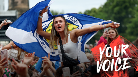 A young woman in a white sleeveless tops is sitting on someone's shoulders, above a large crowd of people. She is waving a Scotland flag. 